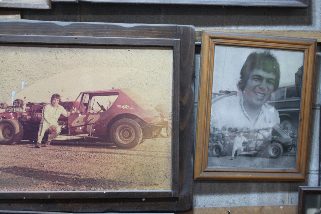 Two framed photographs on a wall, one showing a man posing next to a modified race car, and another portrait of the same man smiling.