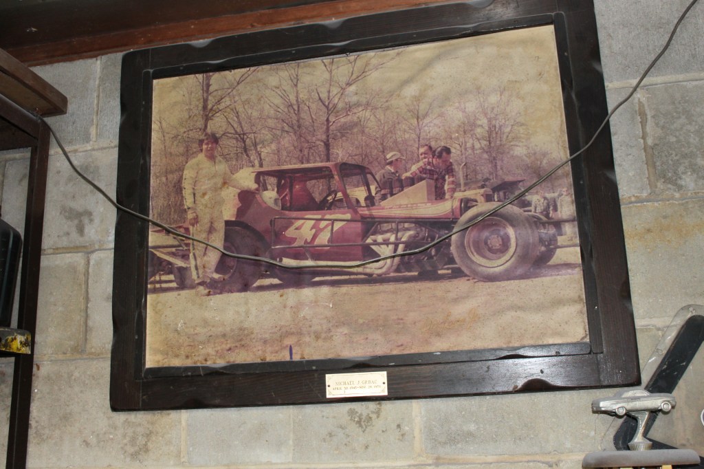 A vintage photograph of a modified race car with four people, including a man in a racing suit, posed next to it, framed and displayed on a garage wall.