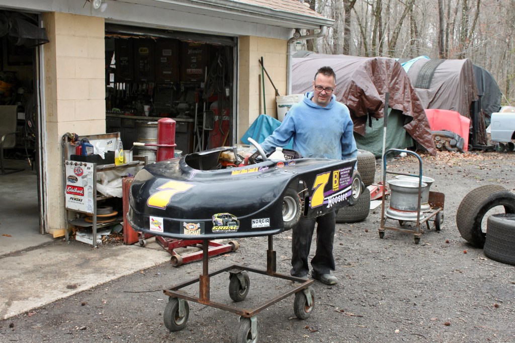 Chris Grbac stands beside a kart near a garage, surrounded by tools and racing memorabilia.